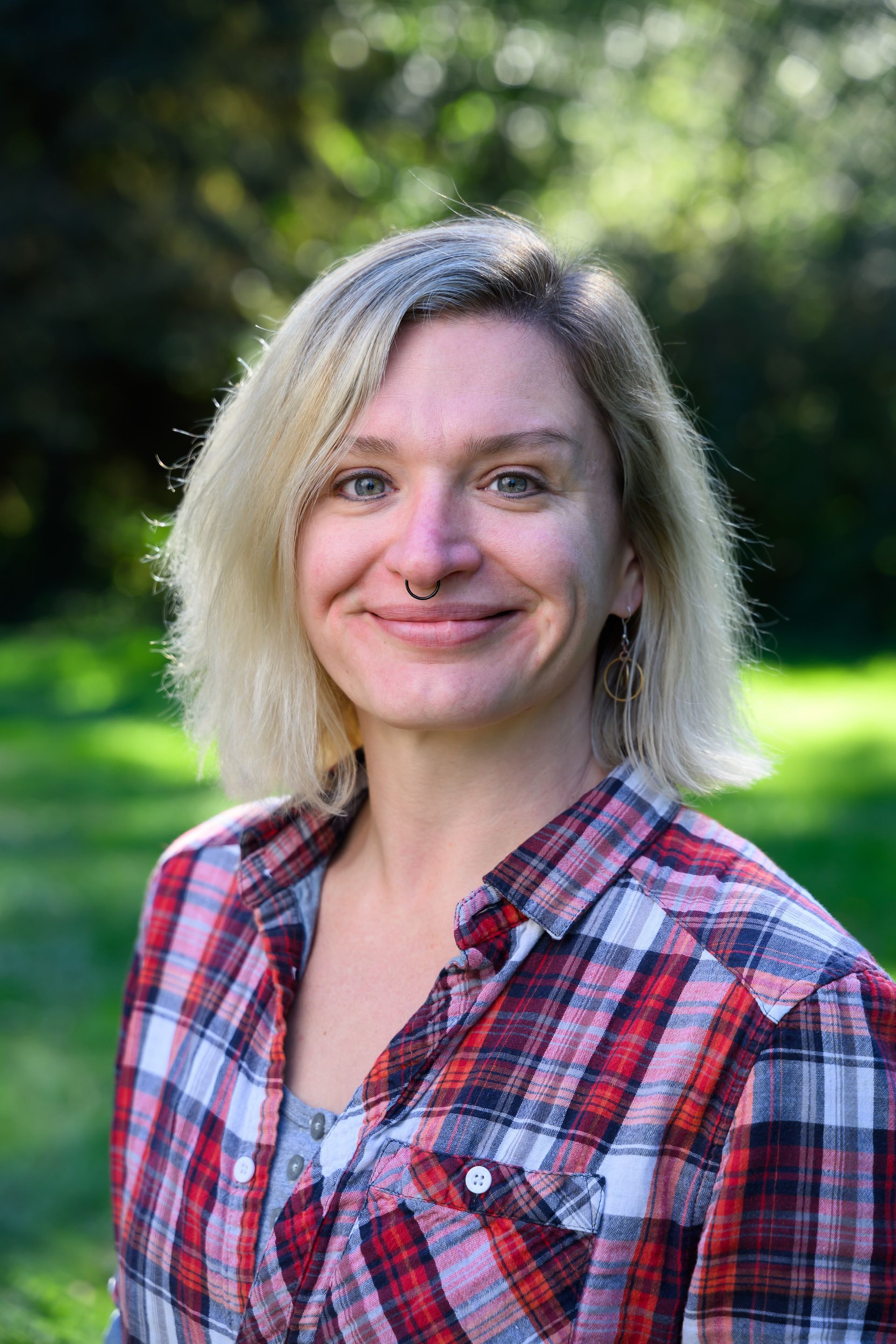 Smiling person in a red plaid shirt standing outdoors with a blurred green background