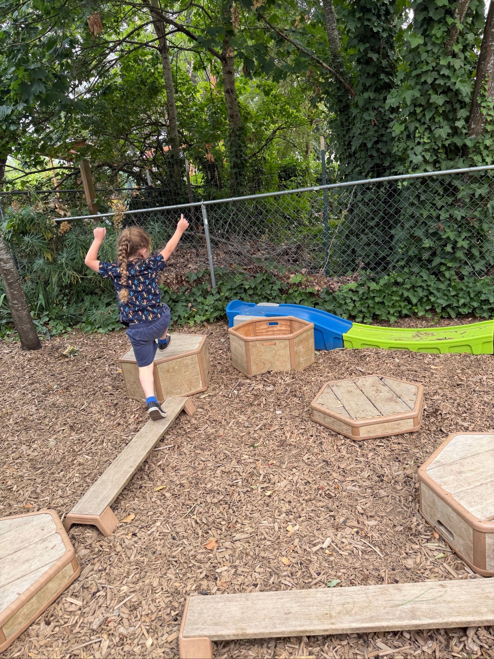 Child balancing on stepping logs in a wooded obstacle course, with a rope line and wood chips below