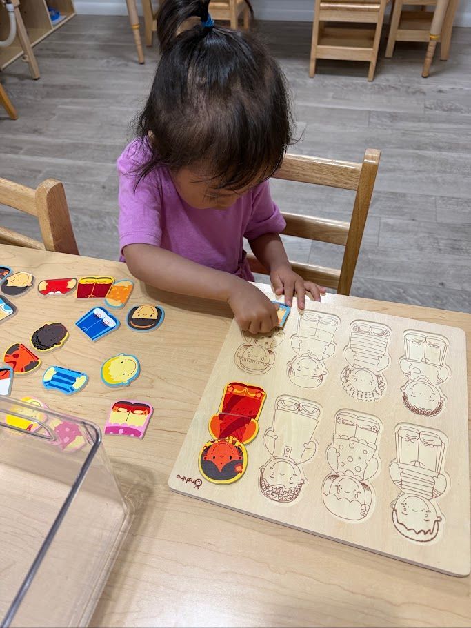 Child in purple shirt placing colorful puzzle pieces on a wooden table in a classroom.