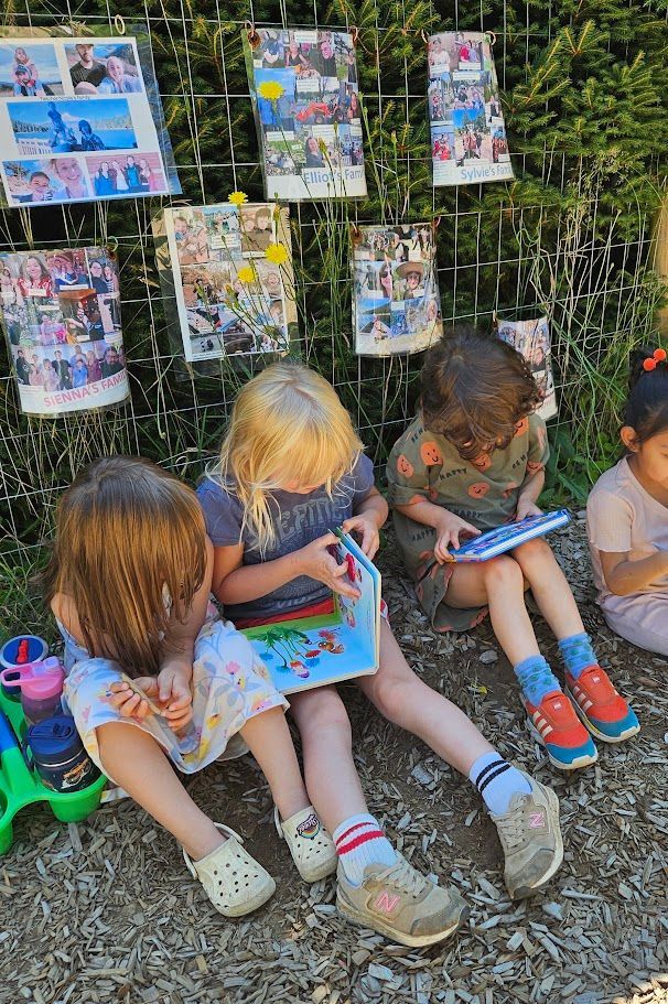 Children seated outdoors, coloring on clipboards beside a fence with hanging artwork.