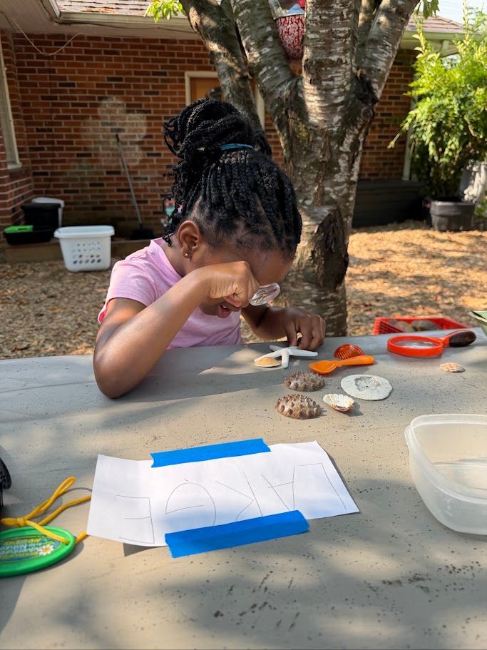 Child painting at a table outdoors under a tree, with art supplies and paper spread out.