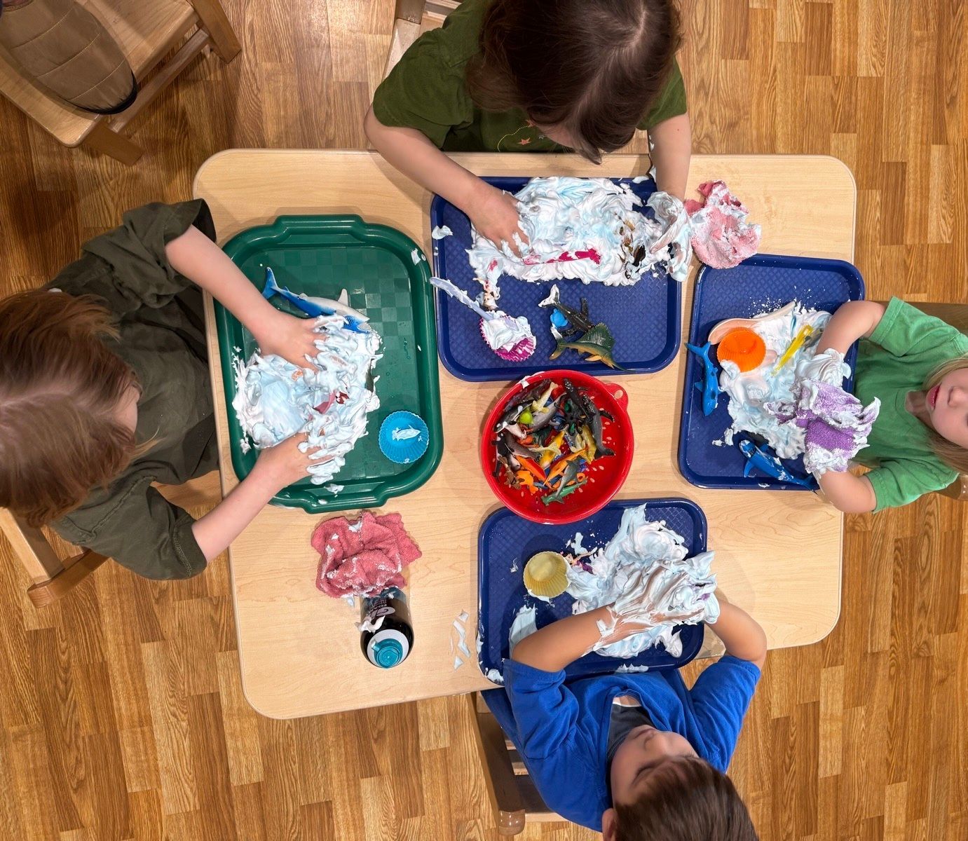 Children playing with shaving cream and colorful objects in trays around a wooden table.