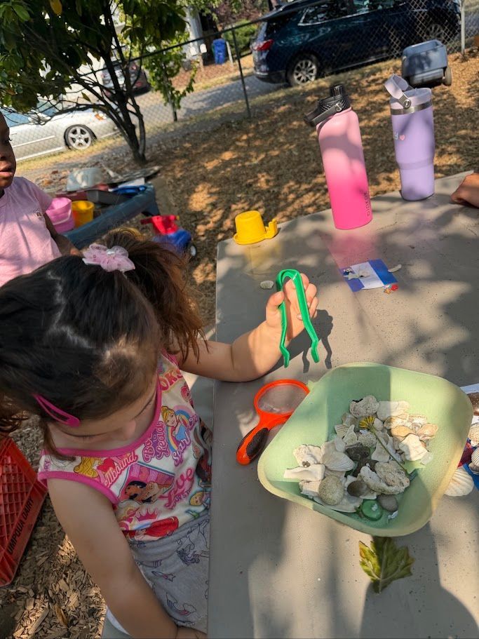 Child playing with toys at a picnic table outdoors, holding a green toy beside a bowl of sand and rocks.