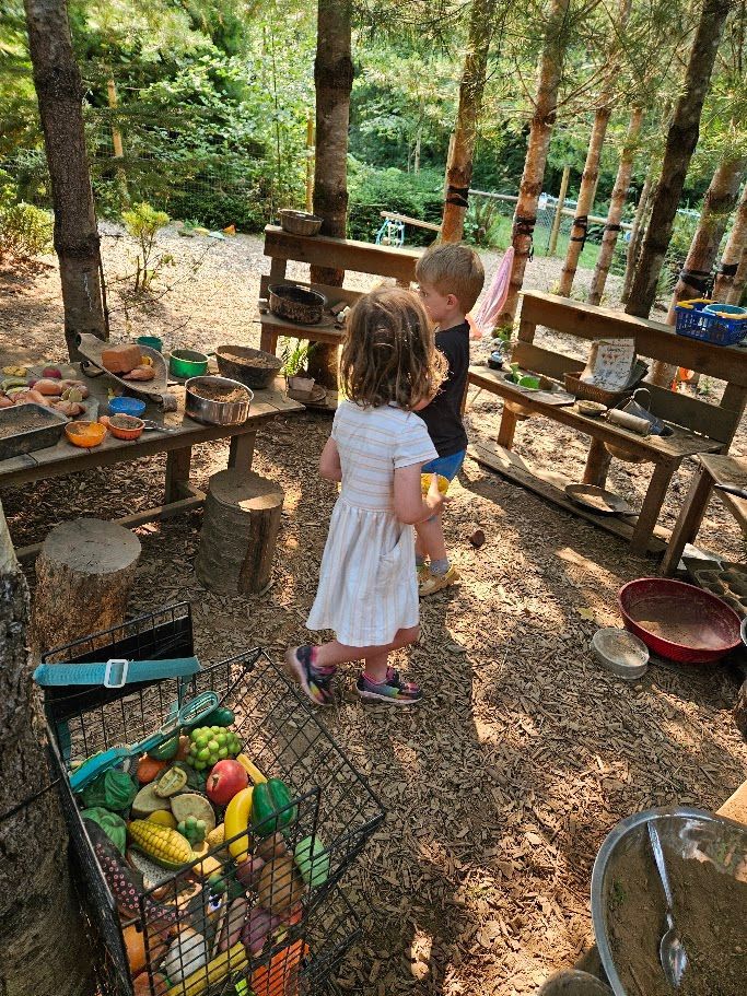 Children playing in a rustic outdoor kitchen with wooden shelves, pots, and baskets among trees.