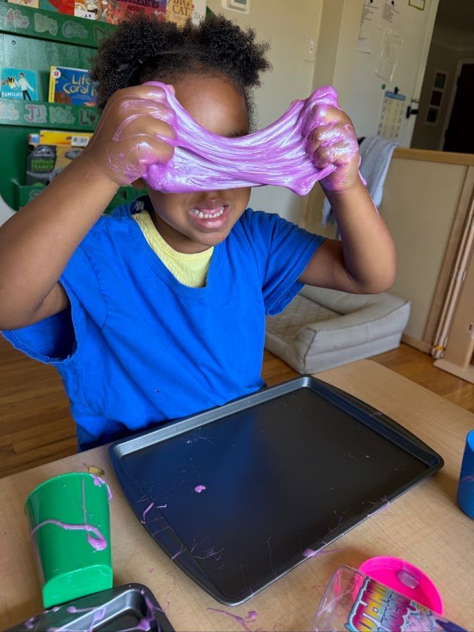 Child stretching pink slime above a tablet at a table, with craft supplies nearby.