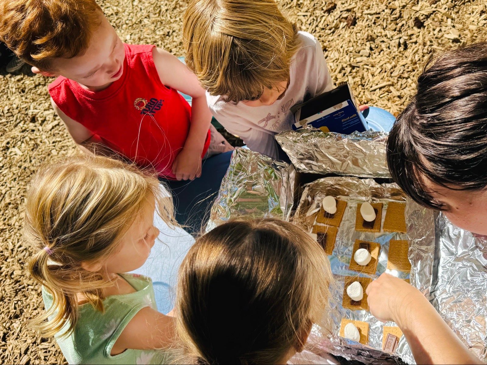 Children gathered around a reflective tray outdoors, exploring water and objects together