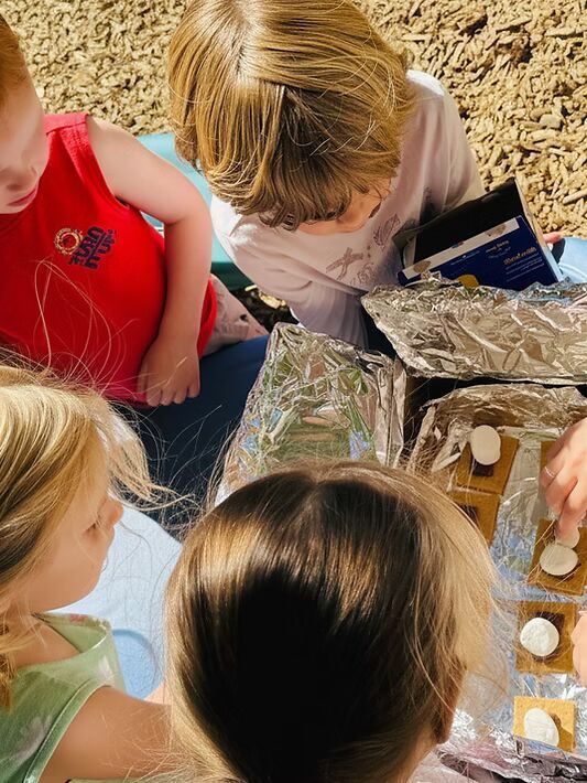 Children gathered around a foil-covered table, mixing ingredients in small containers outdoors.