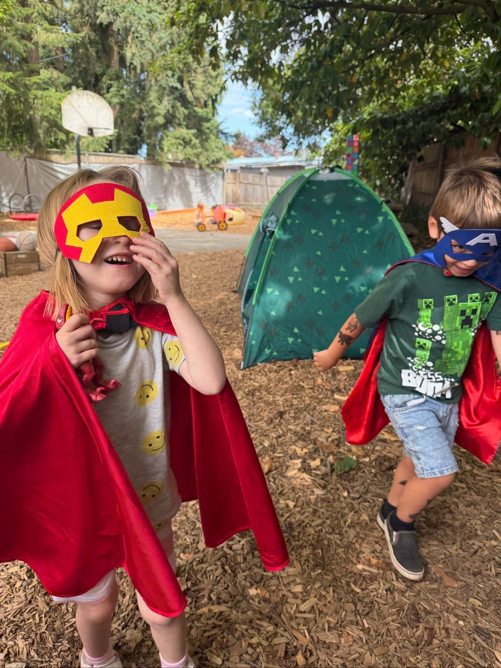 Children in superhero capes playing outdoors near a small green tent on a leaf-covered playground
