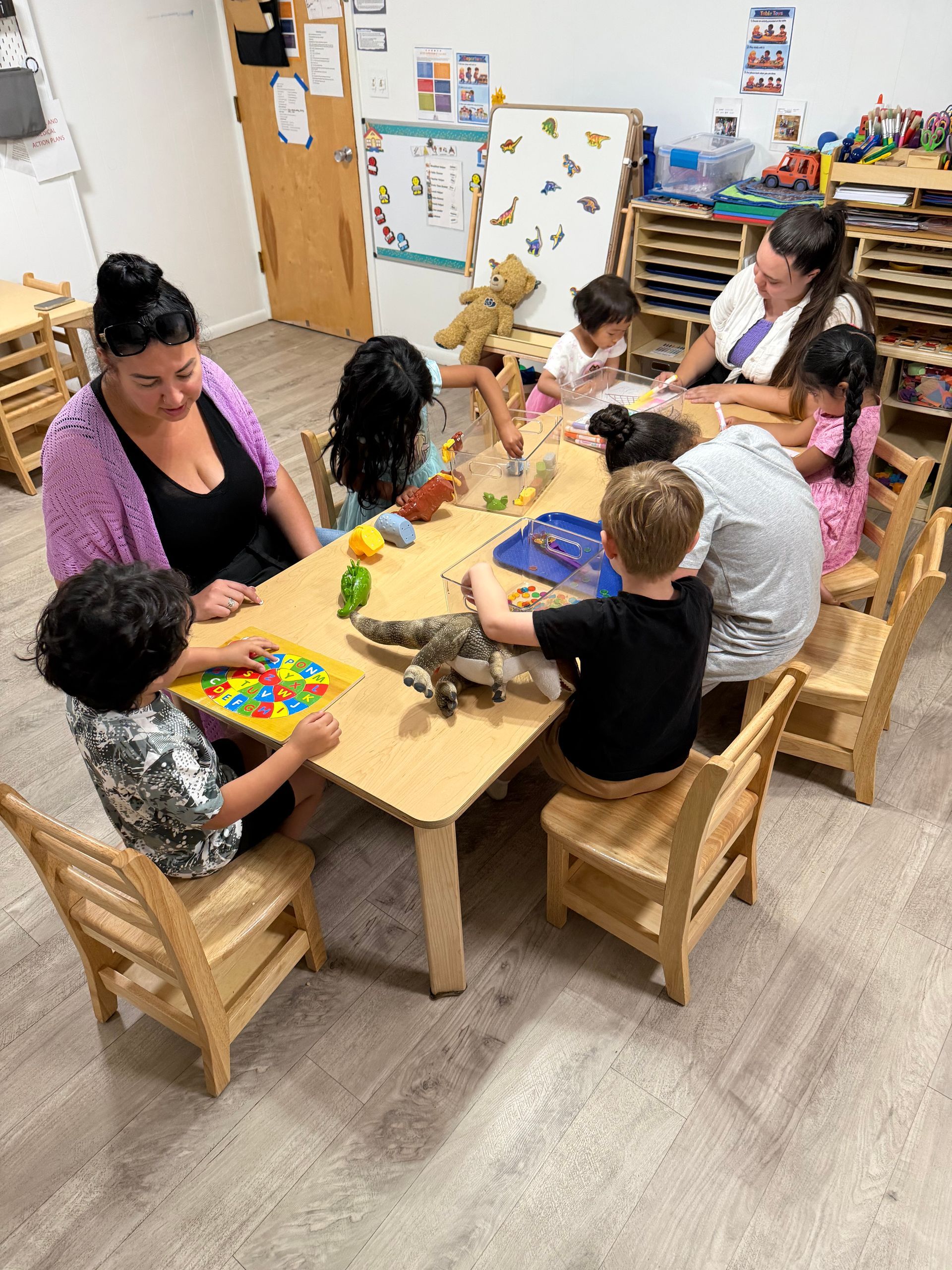 Adults and children sit at a small table doing colorful arts and crafts in a classroom.