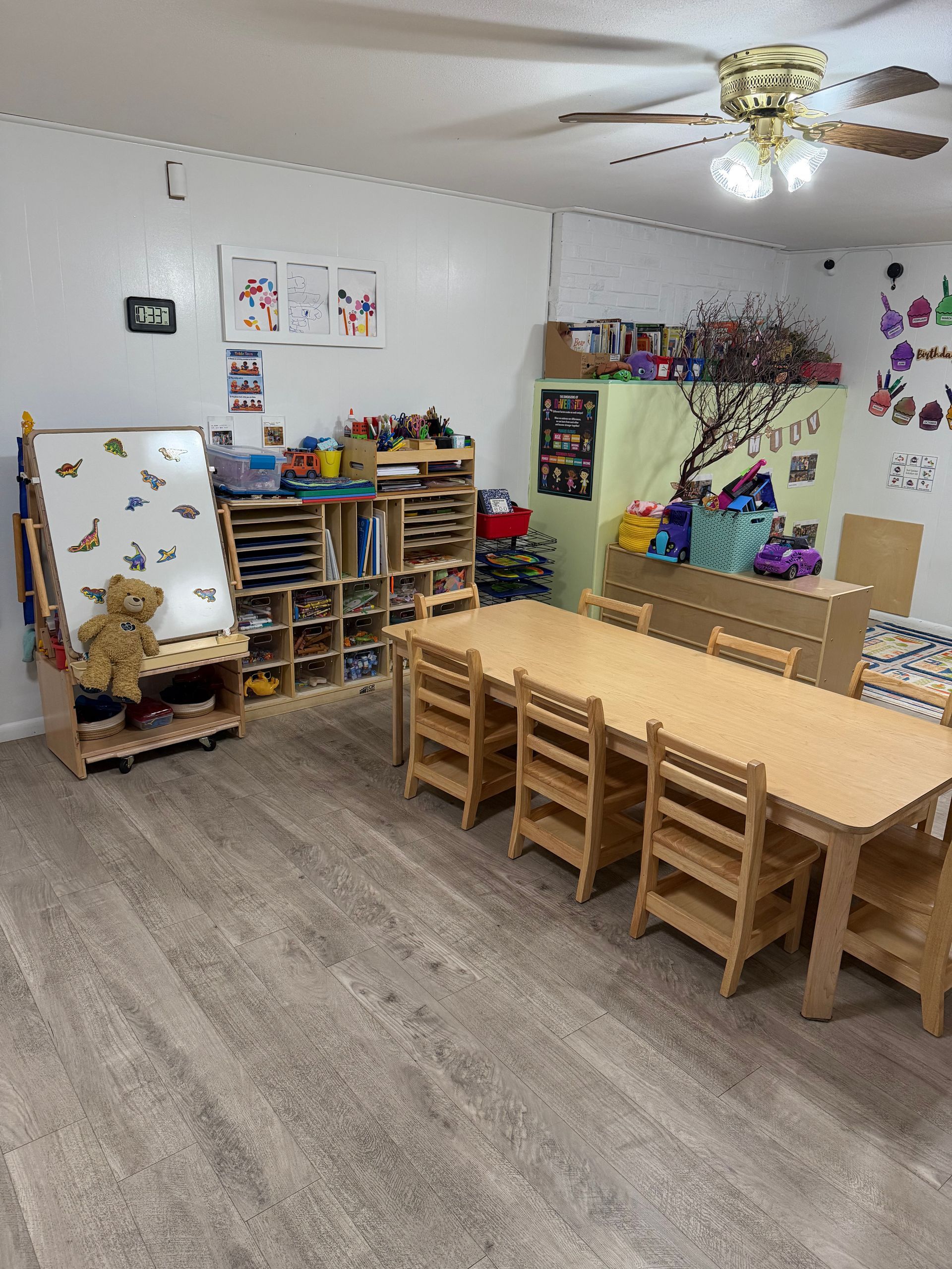 Childcare classroom with wooden tables, shelves, art wall, and play area on carpeted floor