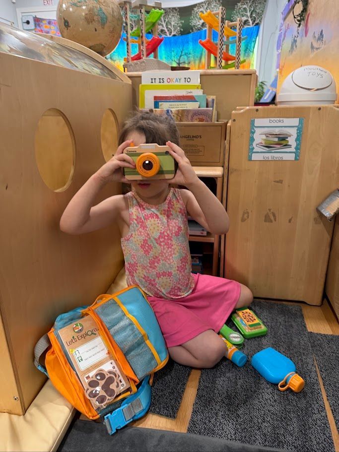 Child sitting in a playroom, holding a toy camera, with toy blocks and a play oven nearby.