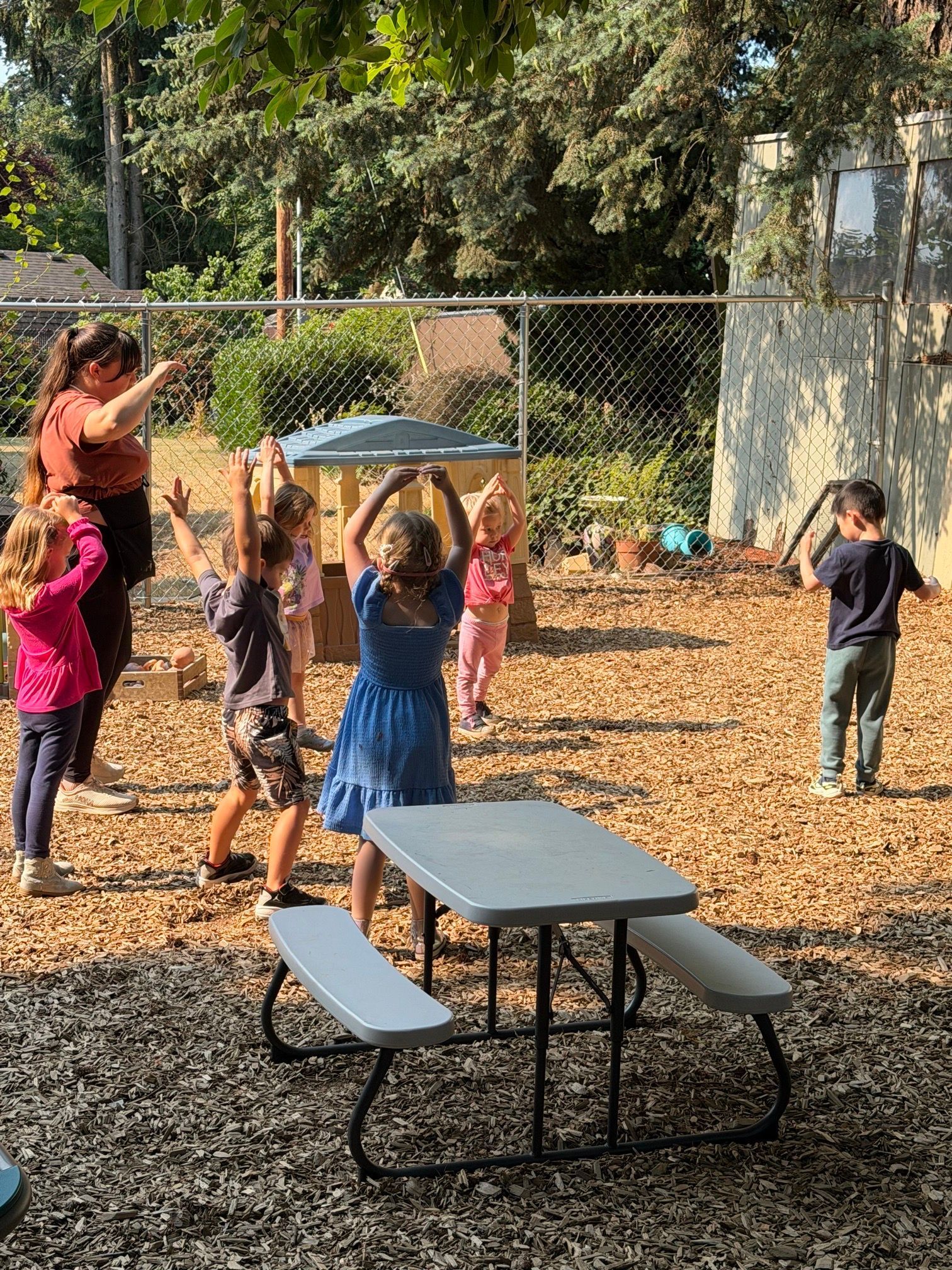 Children playing outdoors near a picnic table, with one child holding a tray overhead under a fence-lined tree.
