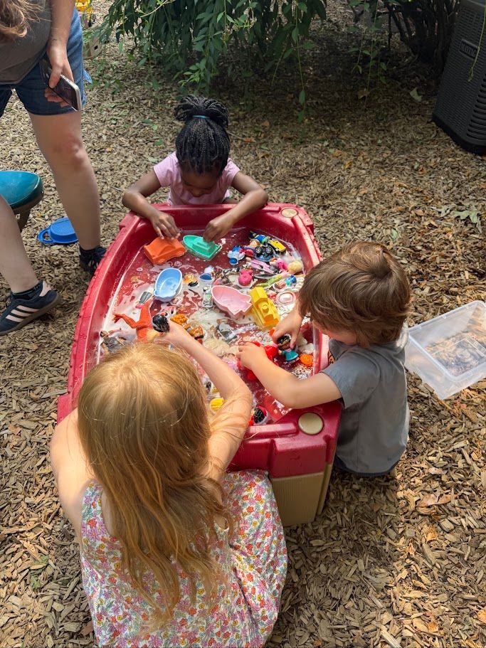 Children playing with toys in a red tub outdoors on a leaf-covered ground