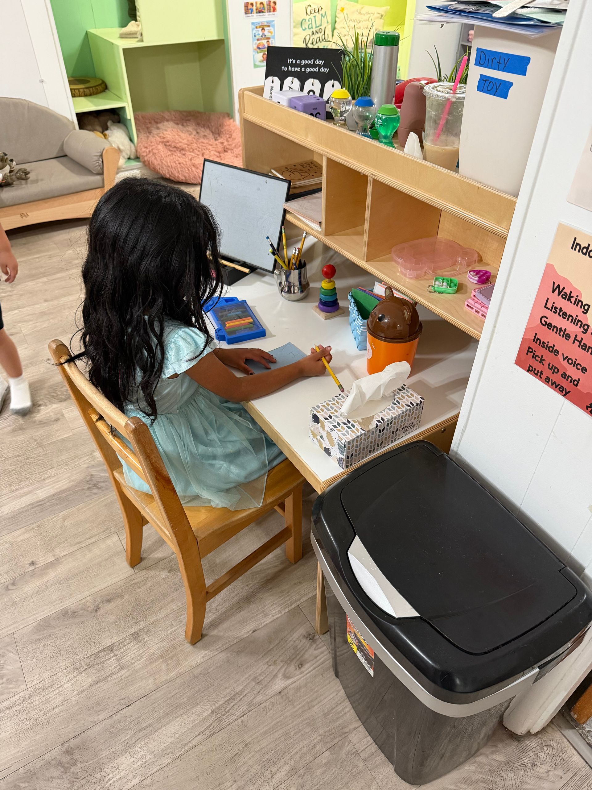 Child sitting at a desk using a laptop in a classroom corner with shelves and art supplies