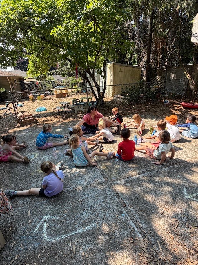 Children sitting in a circle outdoors on gravel, doing an activity under trees.