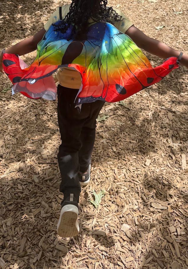 Person walking outdoors on a wooded path, holding a rainbow-colored tie-dye skirt or cape.