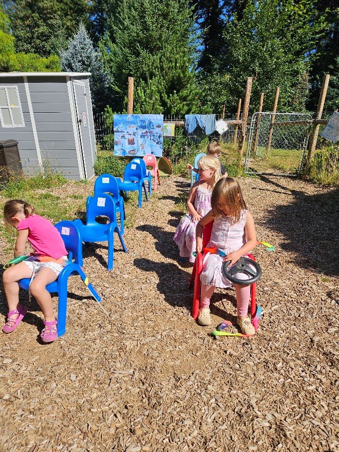 Children sitting on blue chairs in a sunny outdoor yard with a wooden fence and laundry line nearby