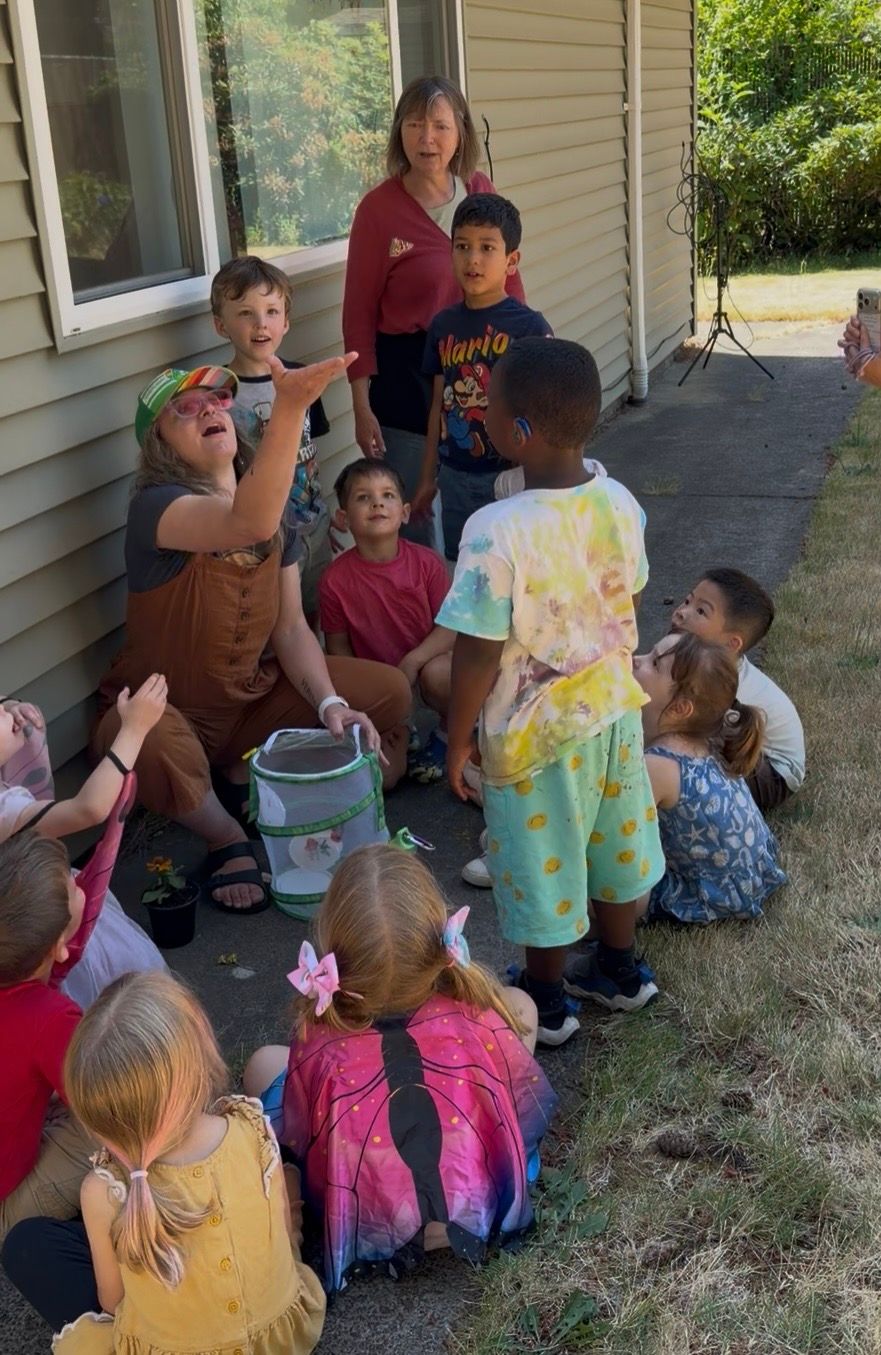 Children gathered on a porch and lawn, sitting around an adult with buckets and toys nearby.