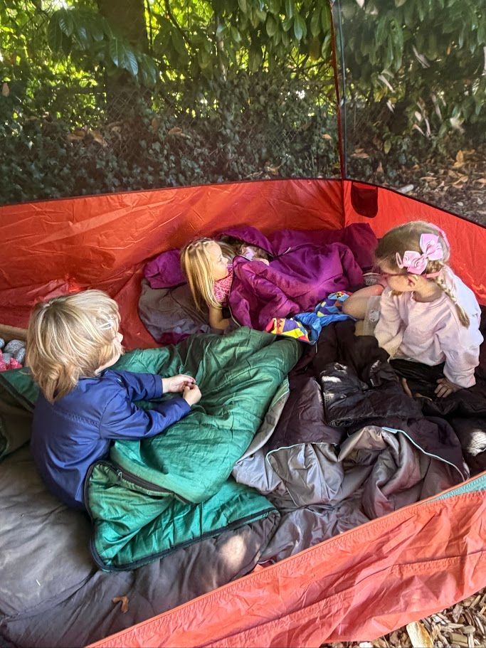 Three children resting in sleeping bags inside a red tent in a wooded campsite.