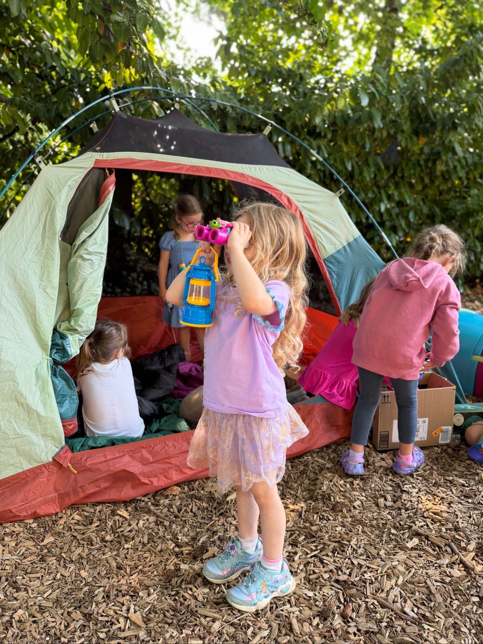 Children playing around a red and green tent under leafy trees, with wood chips on the ground