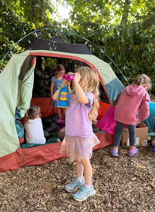 Children gathered around a small camping tent under trees, playing outdoors with toys and backpacks.