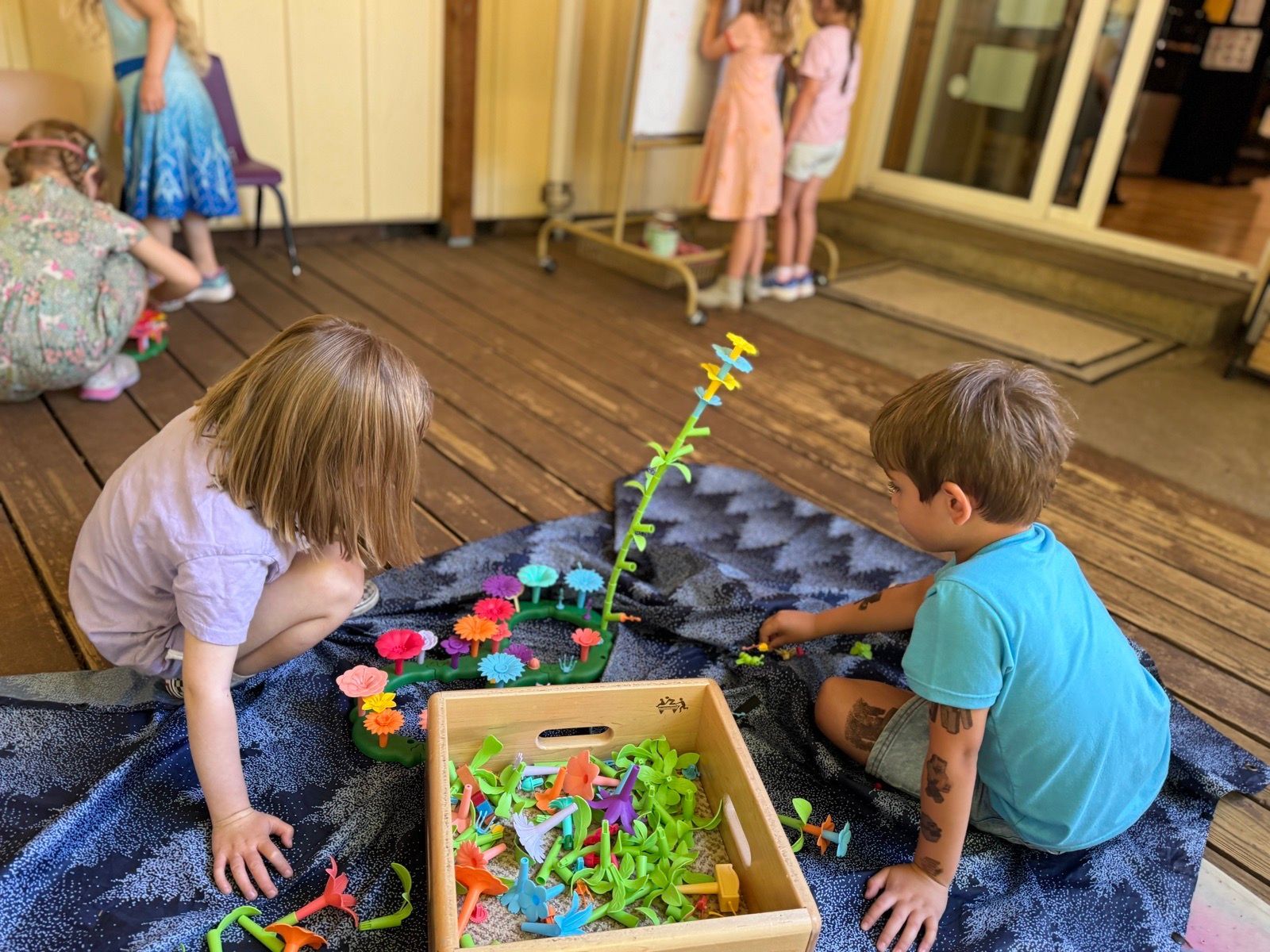 Two children playing with colorful toys on a patio floor beside a box of pieces