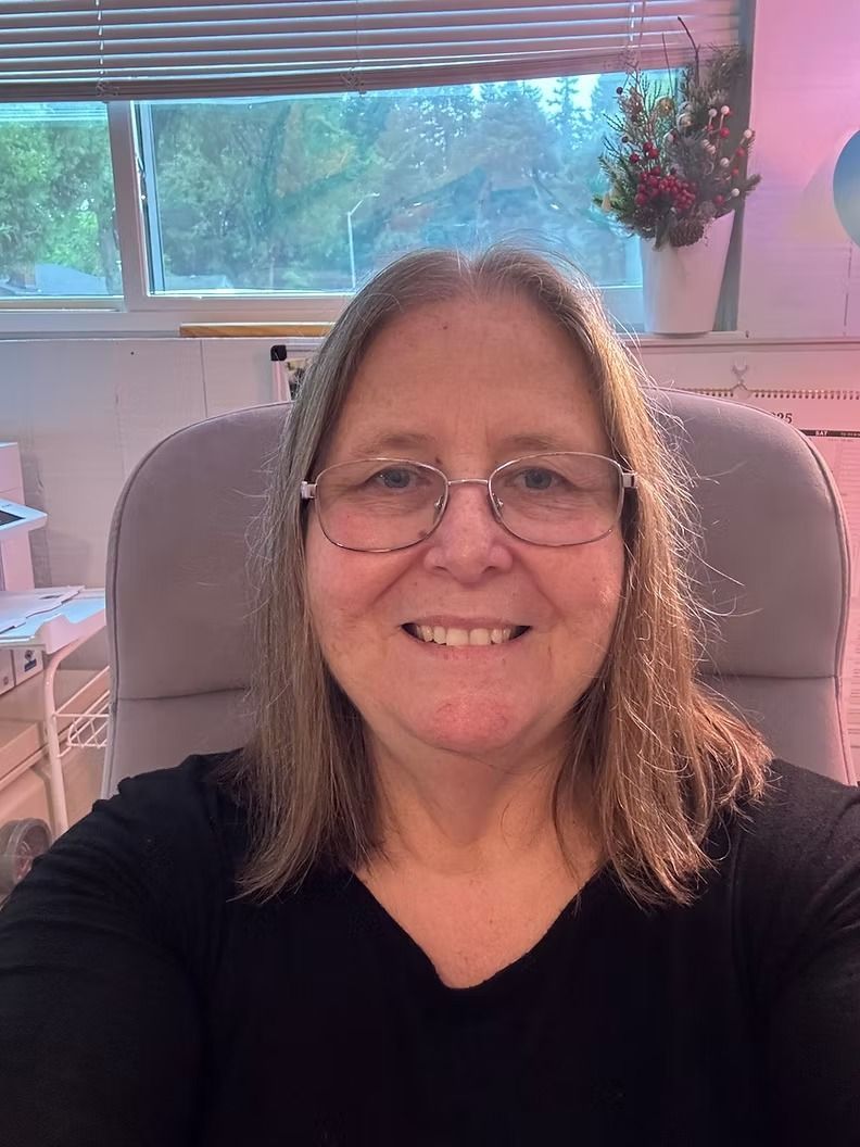 Smiling woman in glasses seated in a home office with a window and hanging plant behind her