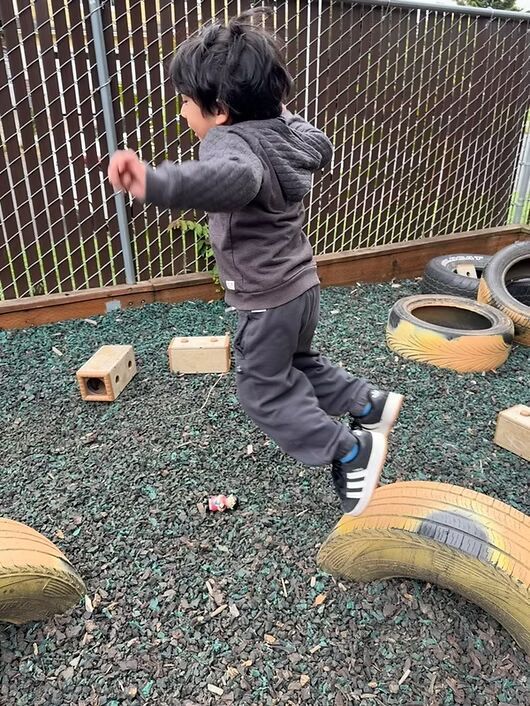 Child jumping between tires in an outdoor play area with gravel and a chain-link fence