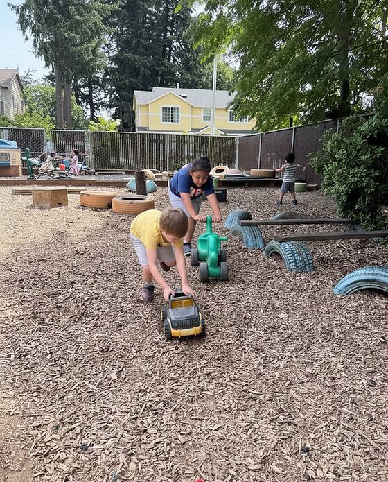 Children pushing toy trucks through wood chips at an outdoor playground.