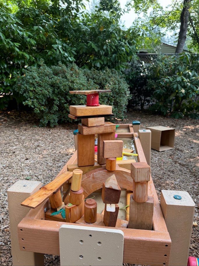 Wooden block tower on a table outdoors, with trees and gravel in the background.