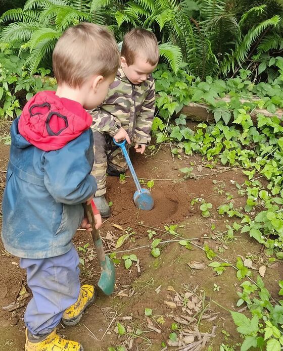 Two children digging in a garden with small shovels beside green plants and ferns