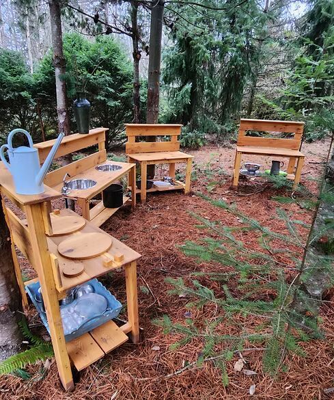 Wooden outdoor toy kitchen set with shelves and a blue watering can in a forest clearing