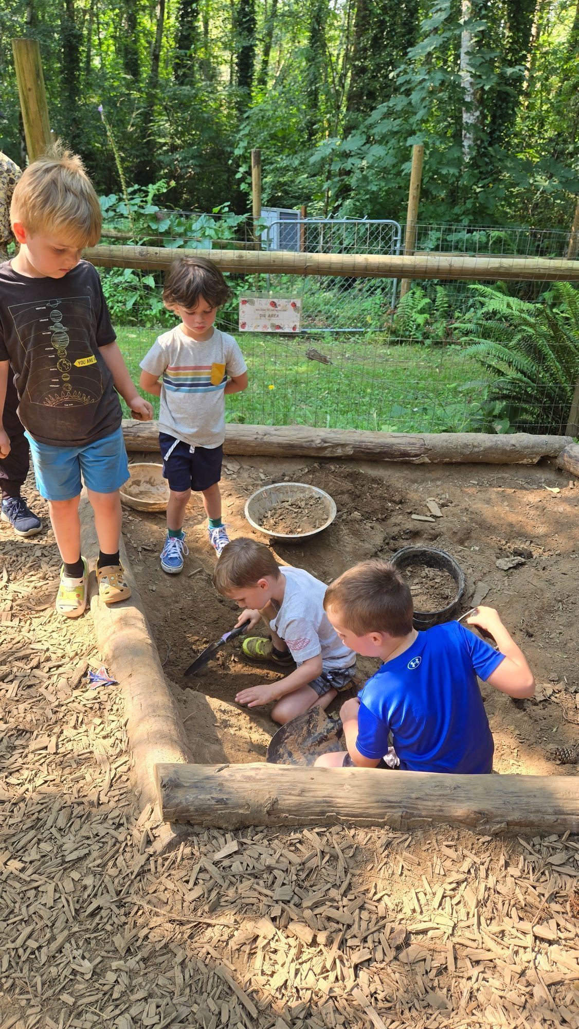 Children playing in a sandy outdoor area near wooden logs and trees