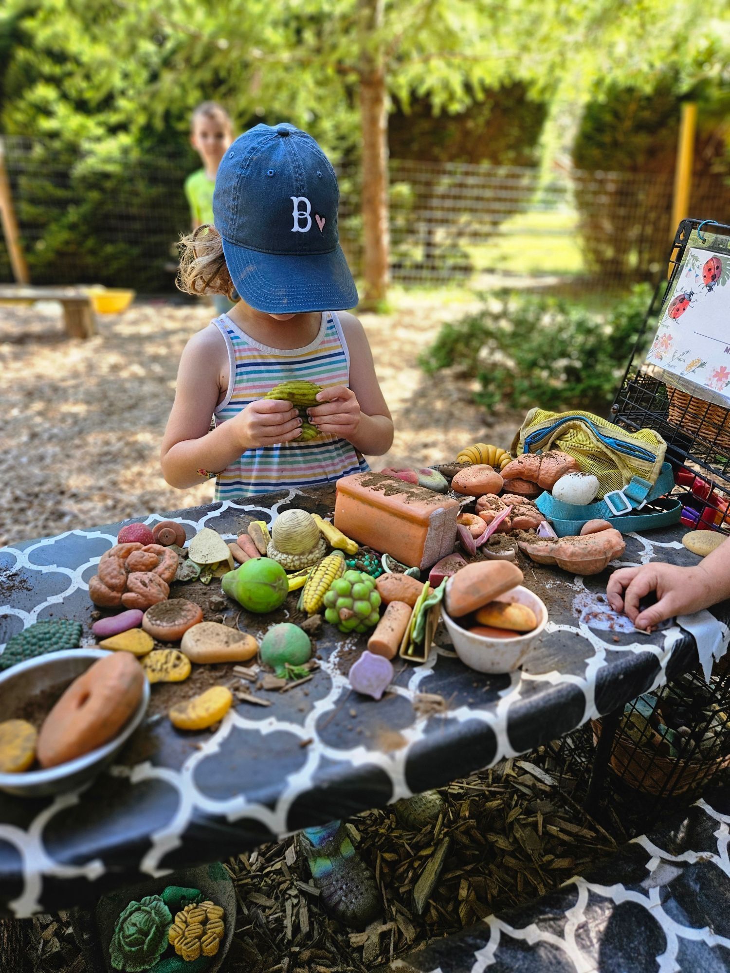 Child in blue cap eating outdoors at a table covered with snacks and drinks in a sunny garden