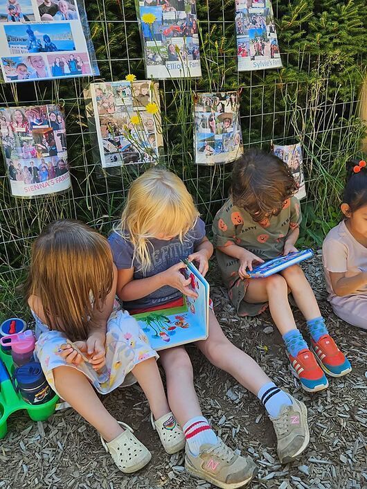 Three children sit on the grass reading colorful books beside a wire fence with photos hanging behind them