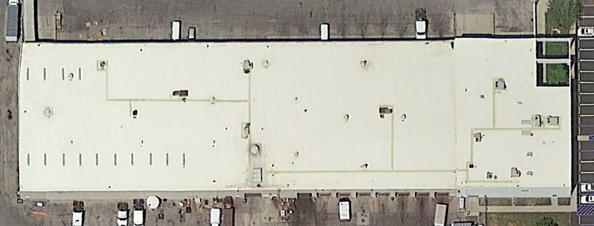 Aerial view of a large, beige industrial building with vents, parking, and green patches.