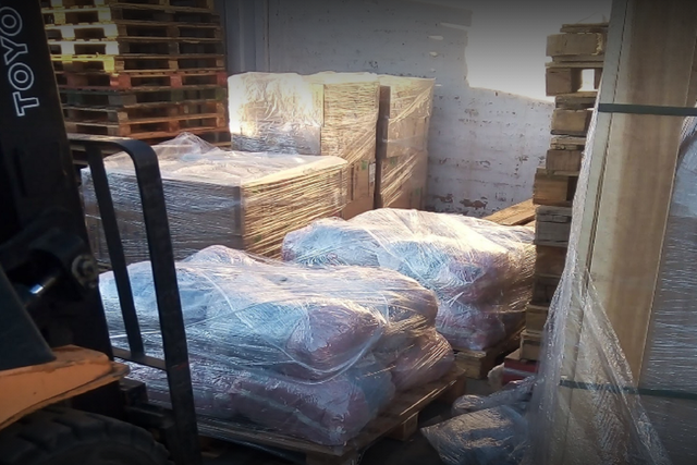 Palés de productos envueltos en plástico retráctil apilados en un almacén junto a una carretilla elevadora.