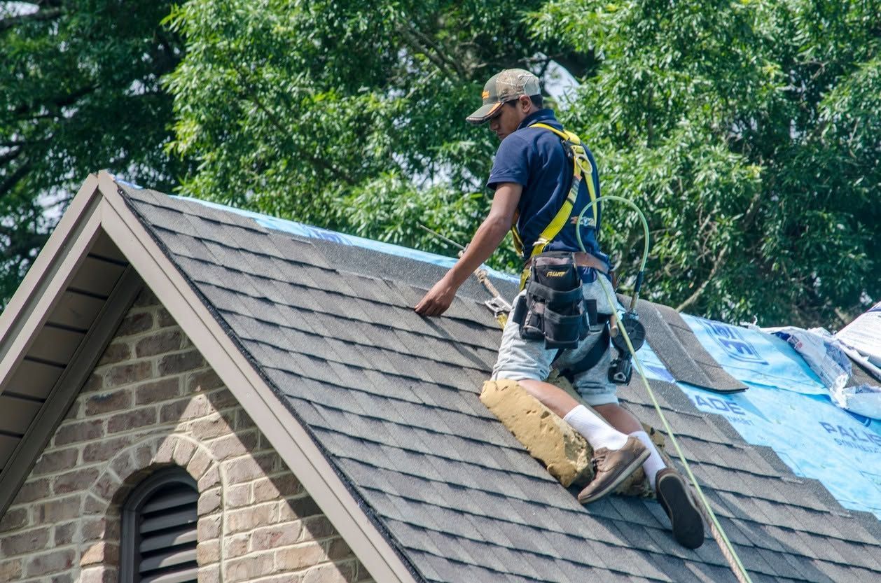 Roofer on a shingled house roof working near blue tarp covering a damaged section