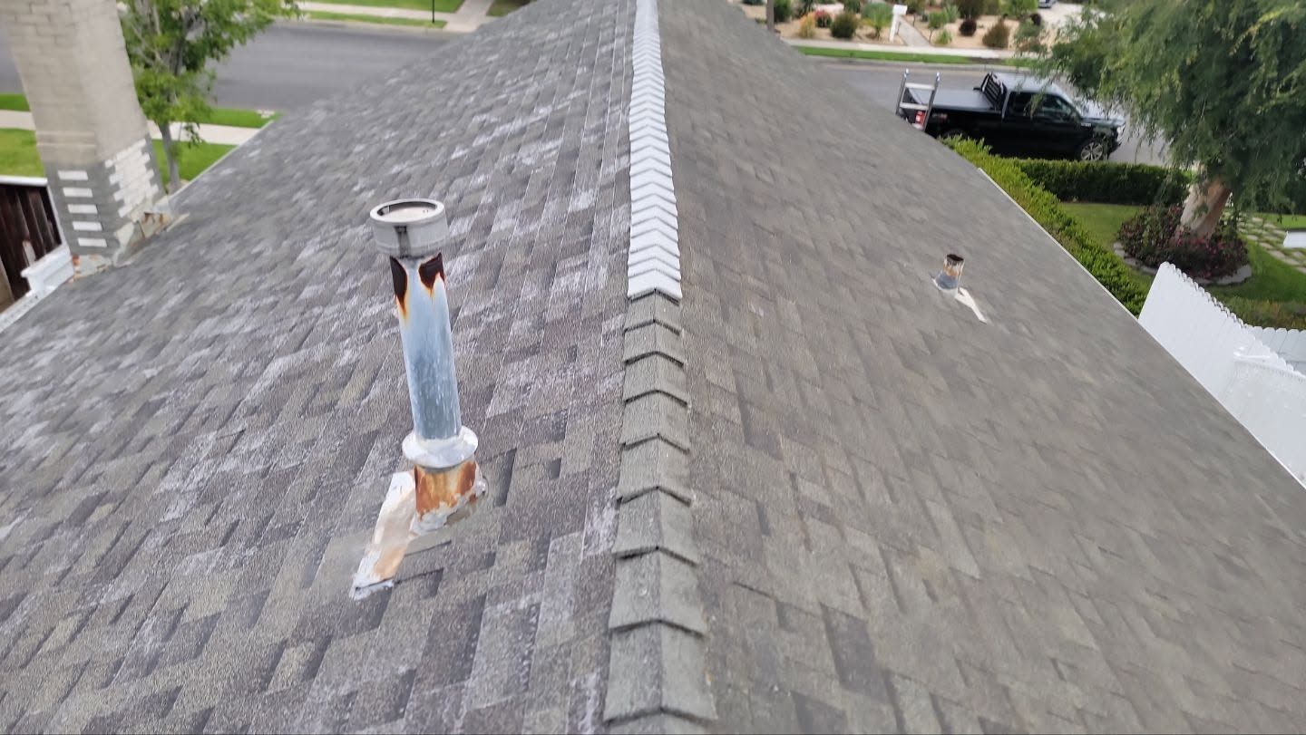 Rooftop view with gray shingles and a metal vent pipe near the roof ridge, with trees and houses beyond