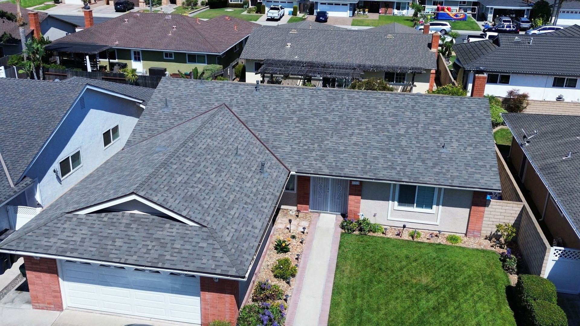 Aerial view of a suburban house with a gray roof, brick exterior, driveway, and green lawn.