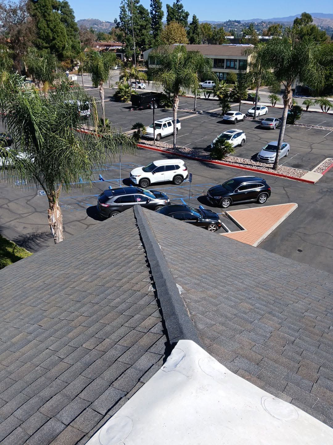 View from a shingled rooftop over a sunny parking lot with several parked cars and trees.
