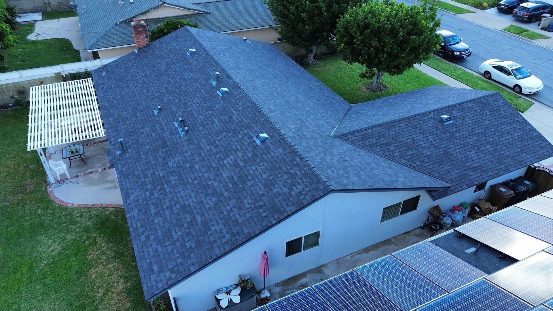 Aerial view of a suburban house with a dark roof, backyard patio, and nearby parked cars.
