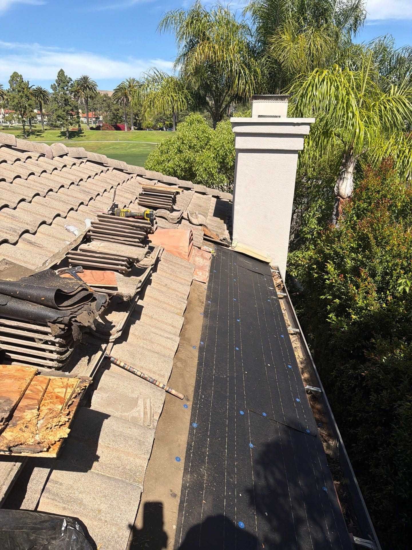 Roof repair beside a white chimney, with stacked tiles and trees in the background