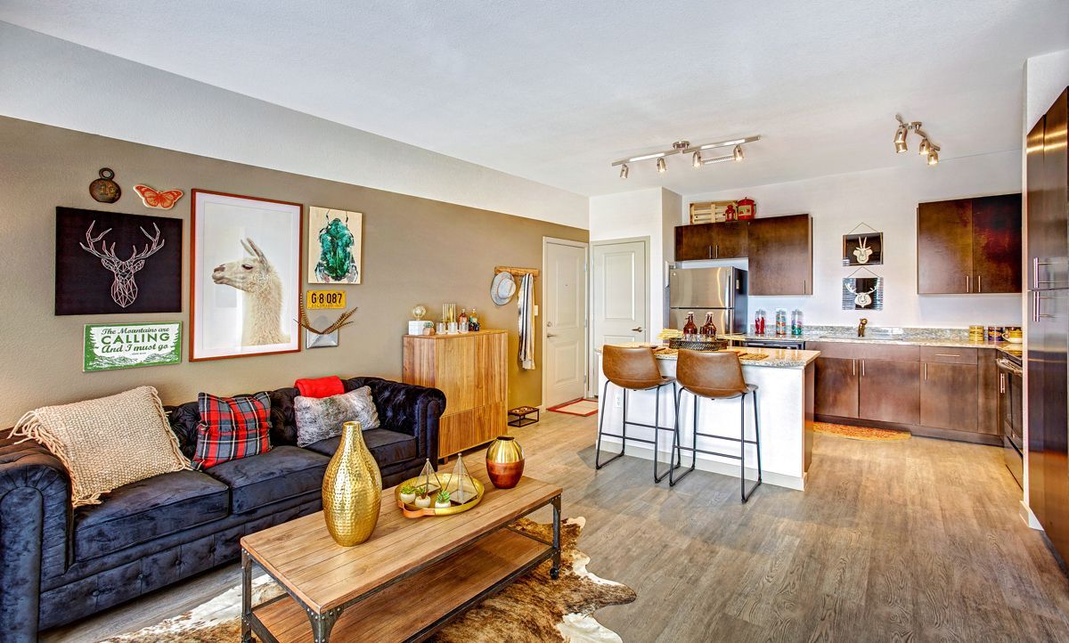 Living room with couch, coffee table, kitchen island, and artwork. Warm neutral tones and modern decor.