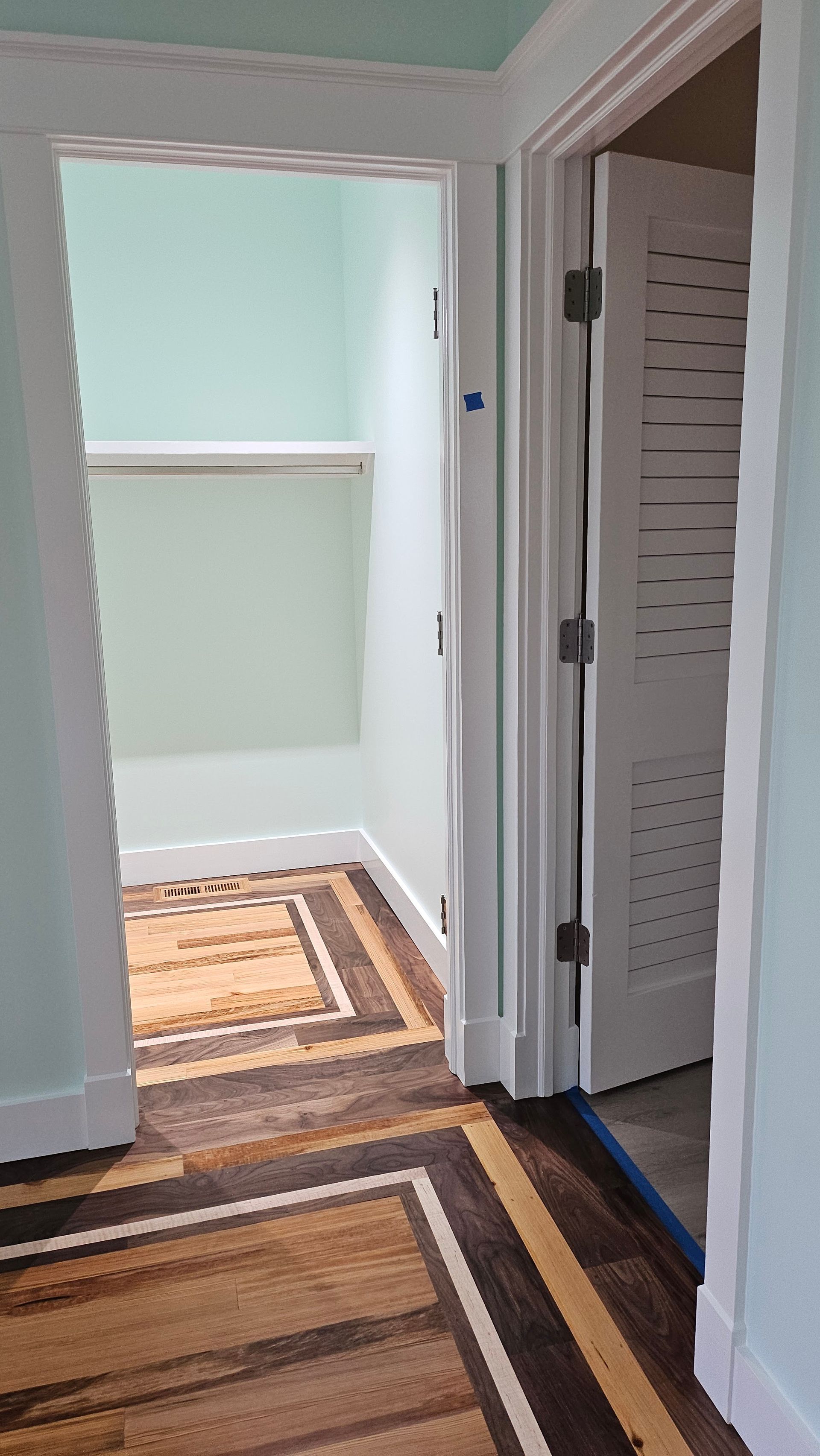 Doorways with patterned hardwood floors. One leads to a small room with a shelf and a mint-green wall. Another is a louvered door.