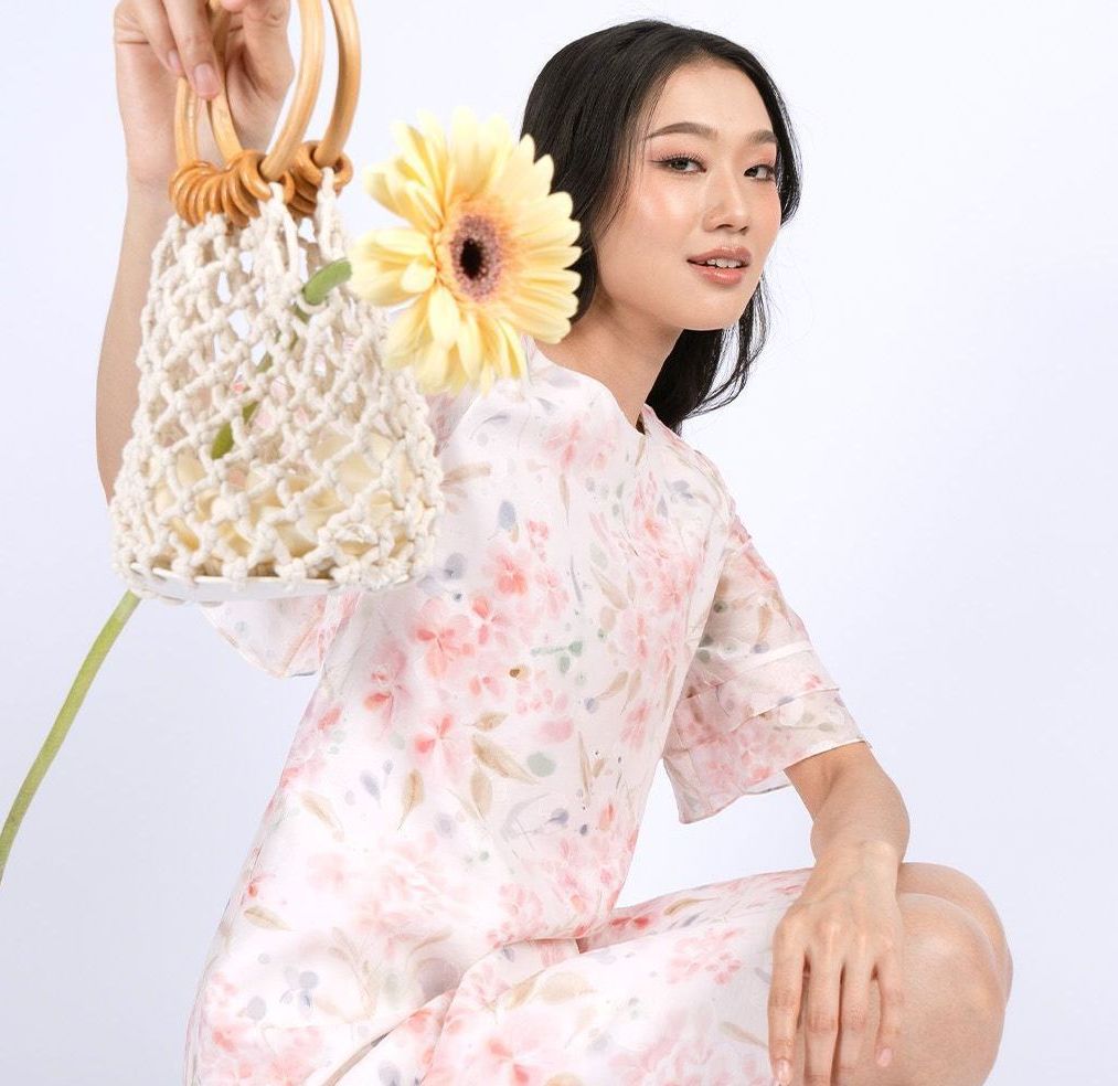 Woman in floral dress holding a crocheted bag and a yellow flower, smiling. White background.