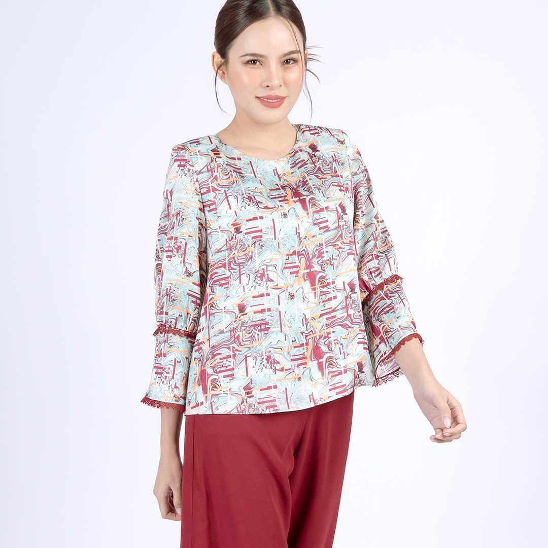 Woman wearing a patterned blouse and red pants smiles. Blouse has 3/4 sleeves and red trim.