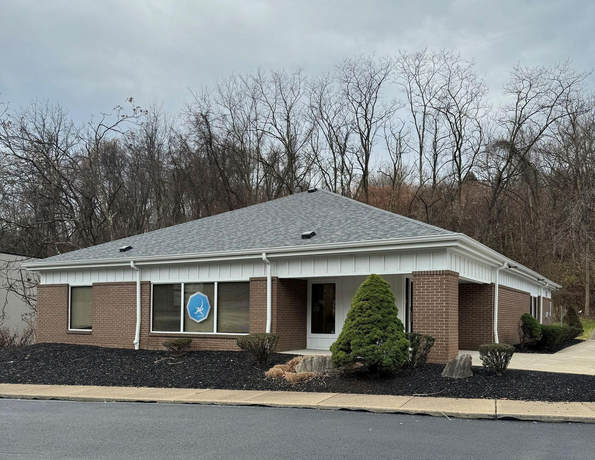 Single-story brick building with a gray roof and a storefront window, surrounded by leafless trees.