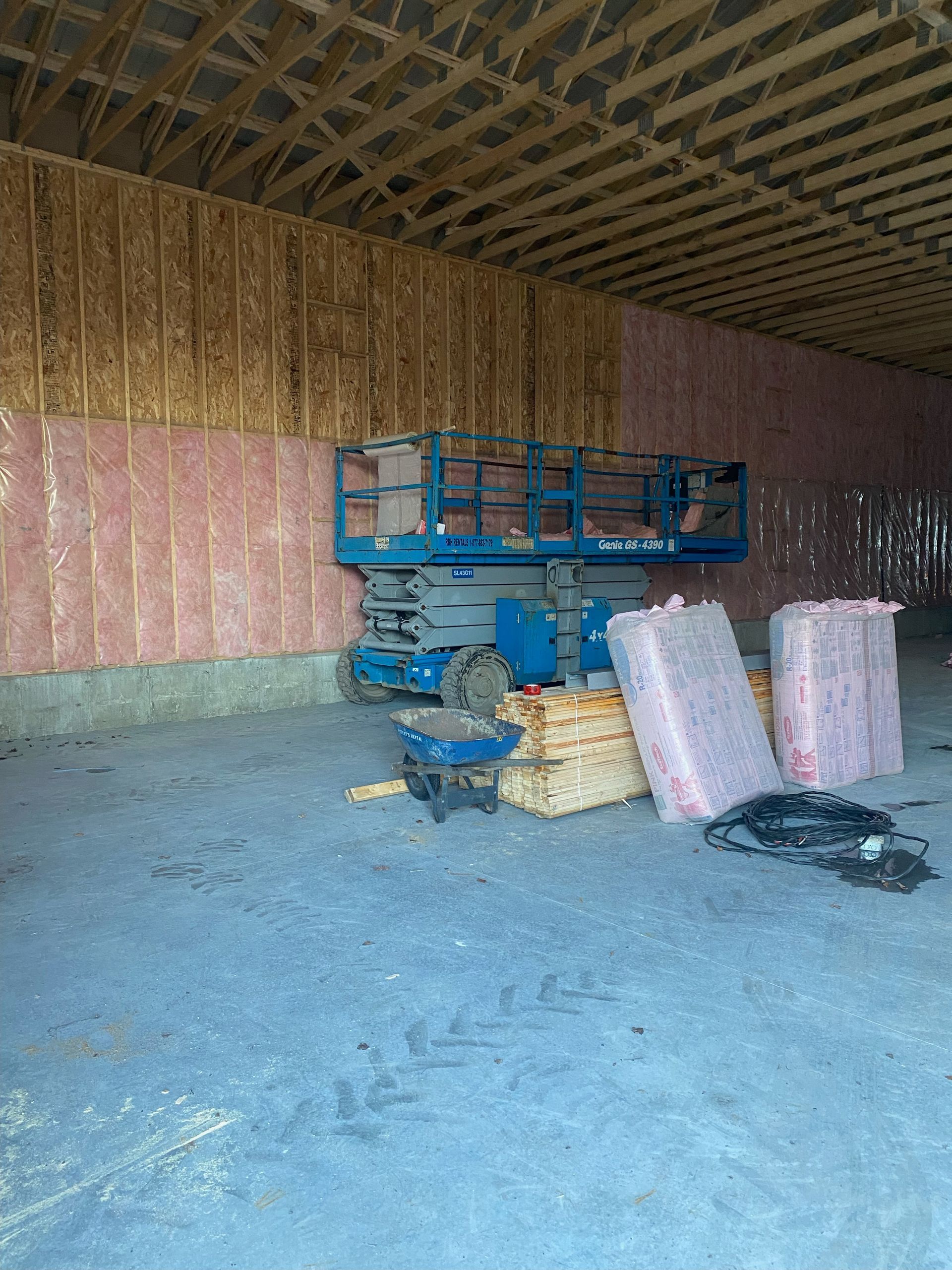 Inside unfinished building with a blue lift, pink insulation, concrete floor, and wood framing.