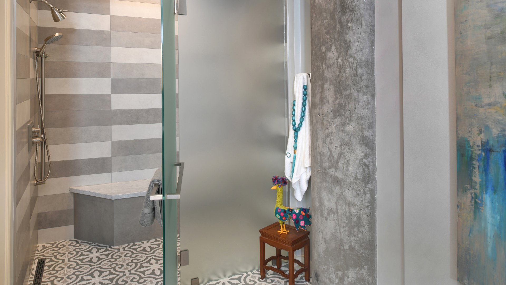 Small bathroom with gray tile walls, glass shower door, toilet, and a stool with hanging clothes.